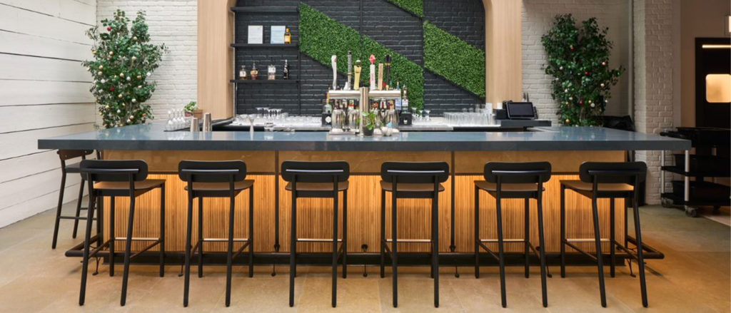 Six Ferdinand Barstools in all black at a bar beneath a skylight. The bar front is lit up from beneath the countertop. Fake hedge greenery is in patches on the wall behind the bar. White painted brick is on either side of the bar.